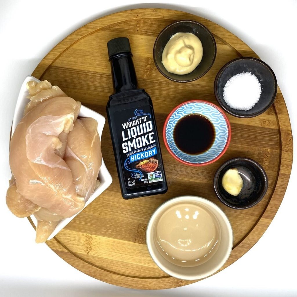 A wood tray with all the ingredients to make the chicken marinade recipe including chicken, liquid smoke, dijon, salt, soy sauce, garlic clove, and wine.