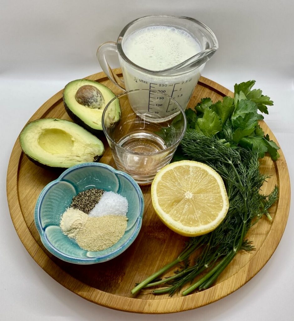 A wood tray with all the ingredients to make the avocado ranch dressing including avocado, buttermilk, fresh herbs, dry seasonings, water and a lemon.