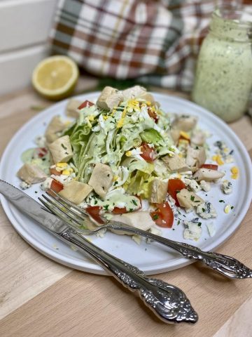 salad on a plate with silverware, dressing, and lemon.