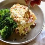 A fork stabbing into a slice of pasta bake alongside steamed broccoli.