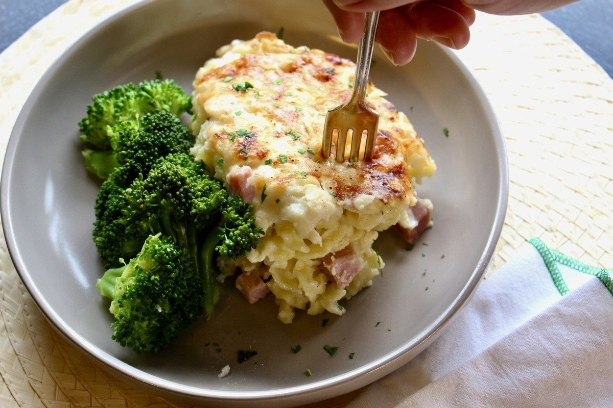 A fork stabbing into a slice of pasta bake alongside steamed broccoli.