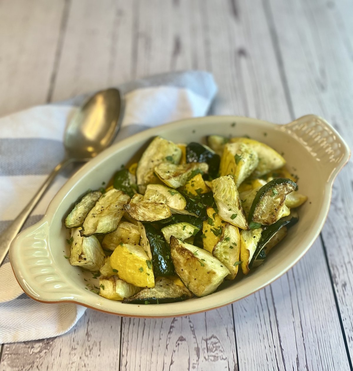 A serving dish filled with air fryer zucchini and yellow squash alongside a spoon and towel.