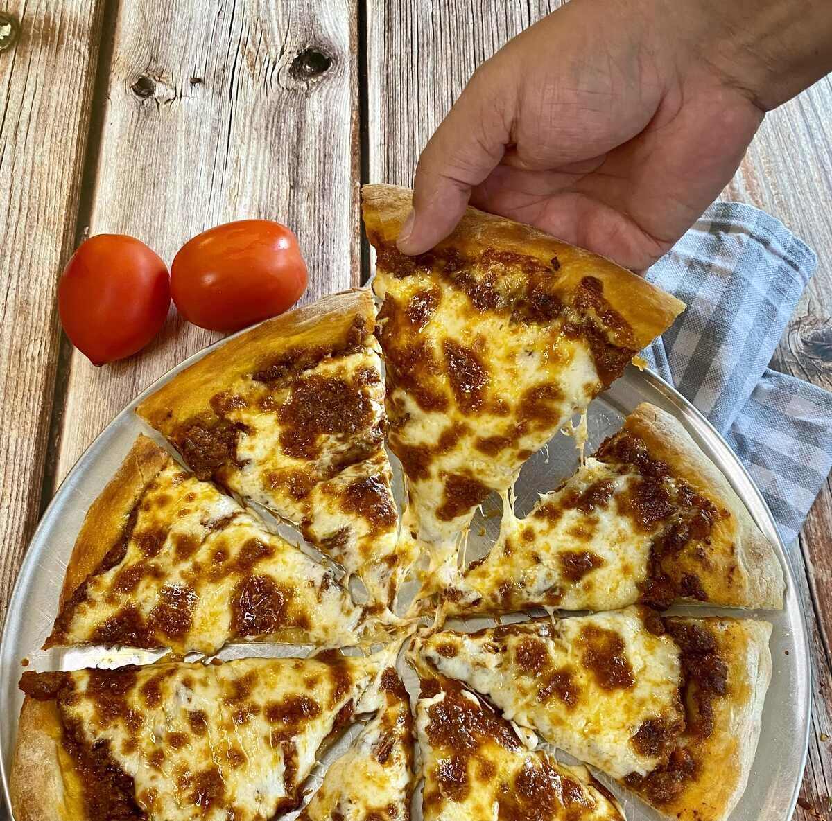 A hand lifting a slice of bolognese pizza off the pizza pan.