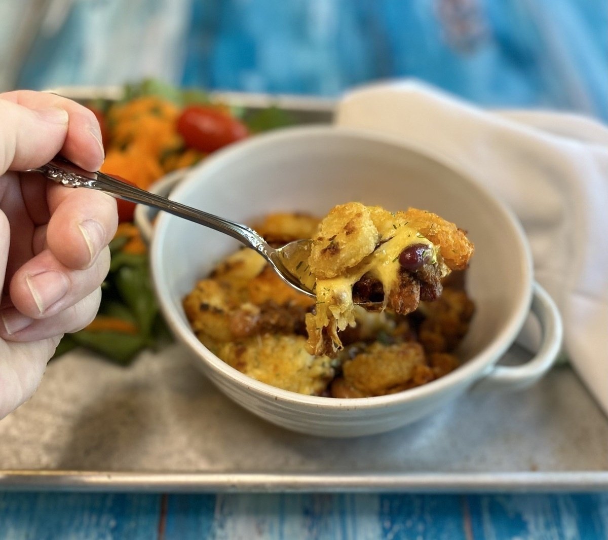 A spoon pulling out a bite of chili tater tot casserole from the bowl with a salad in the background.