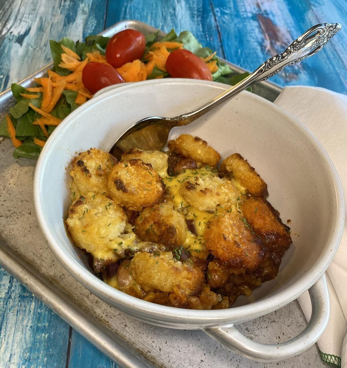 The casserole in a white bowl on a tray with a side salad and a spoon.