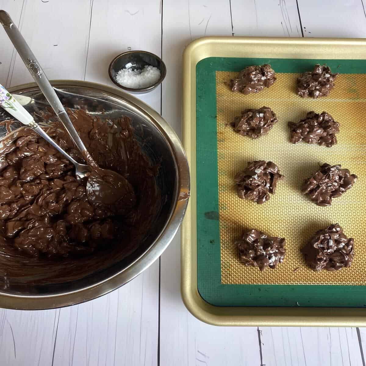 A bowl with cashews and chocolates besides a sheet pan with piles of cashew clusters and maldon flake salt on the side.