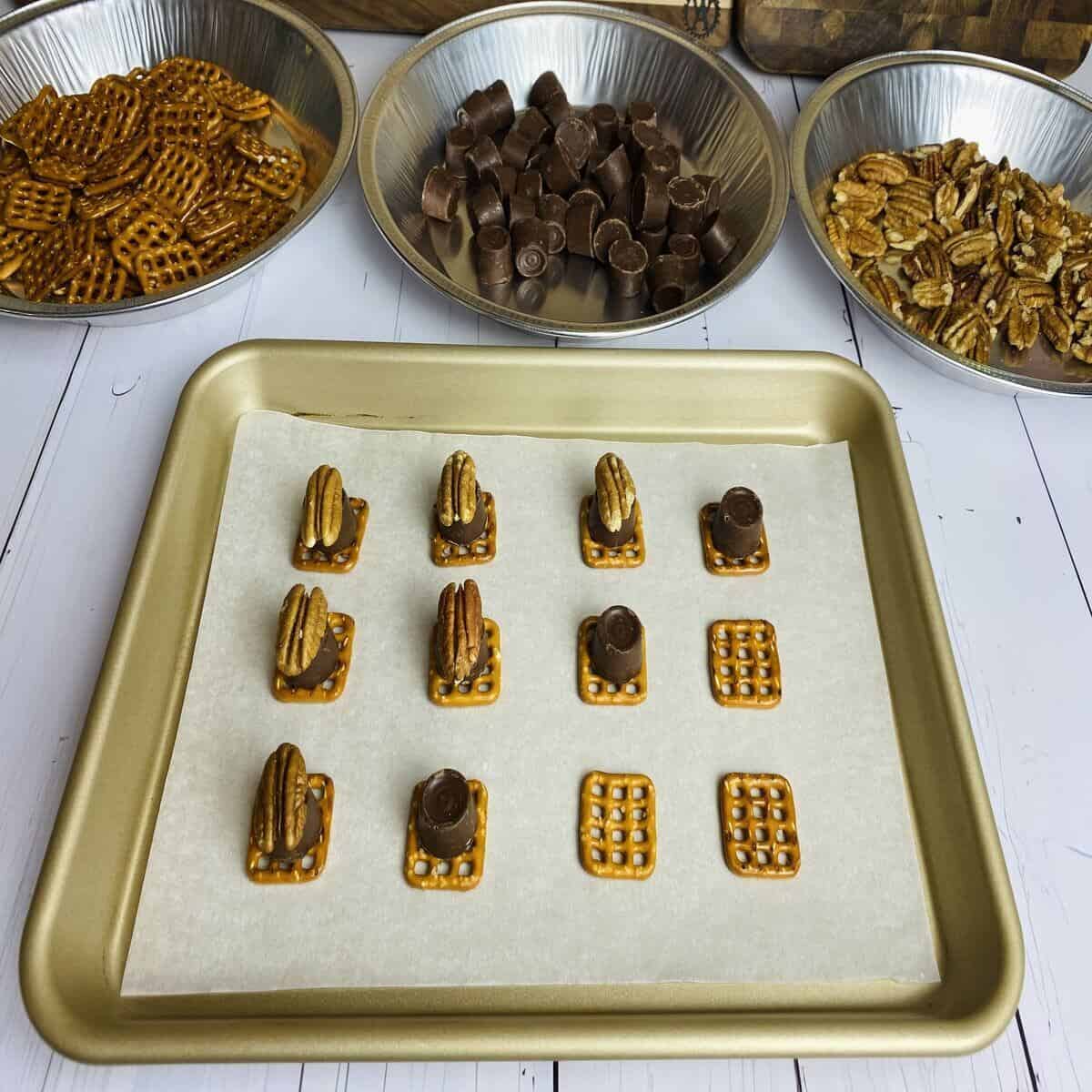 Three pans filled with the ingredients for pecan turtles with a sheet pan in front filled with stacked pretzels, chocolates, and pecans.