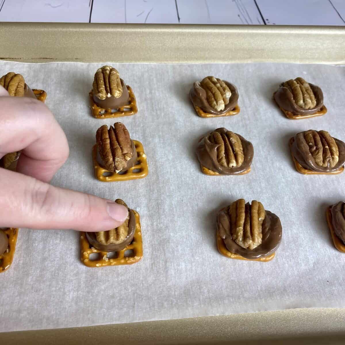 Sheet pan with cooked pecan turtles showing a finger pushing the pecan into the warm chocolate candy.