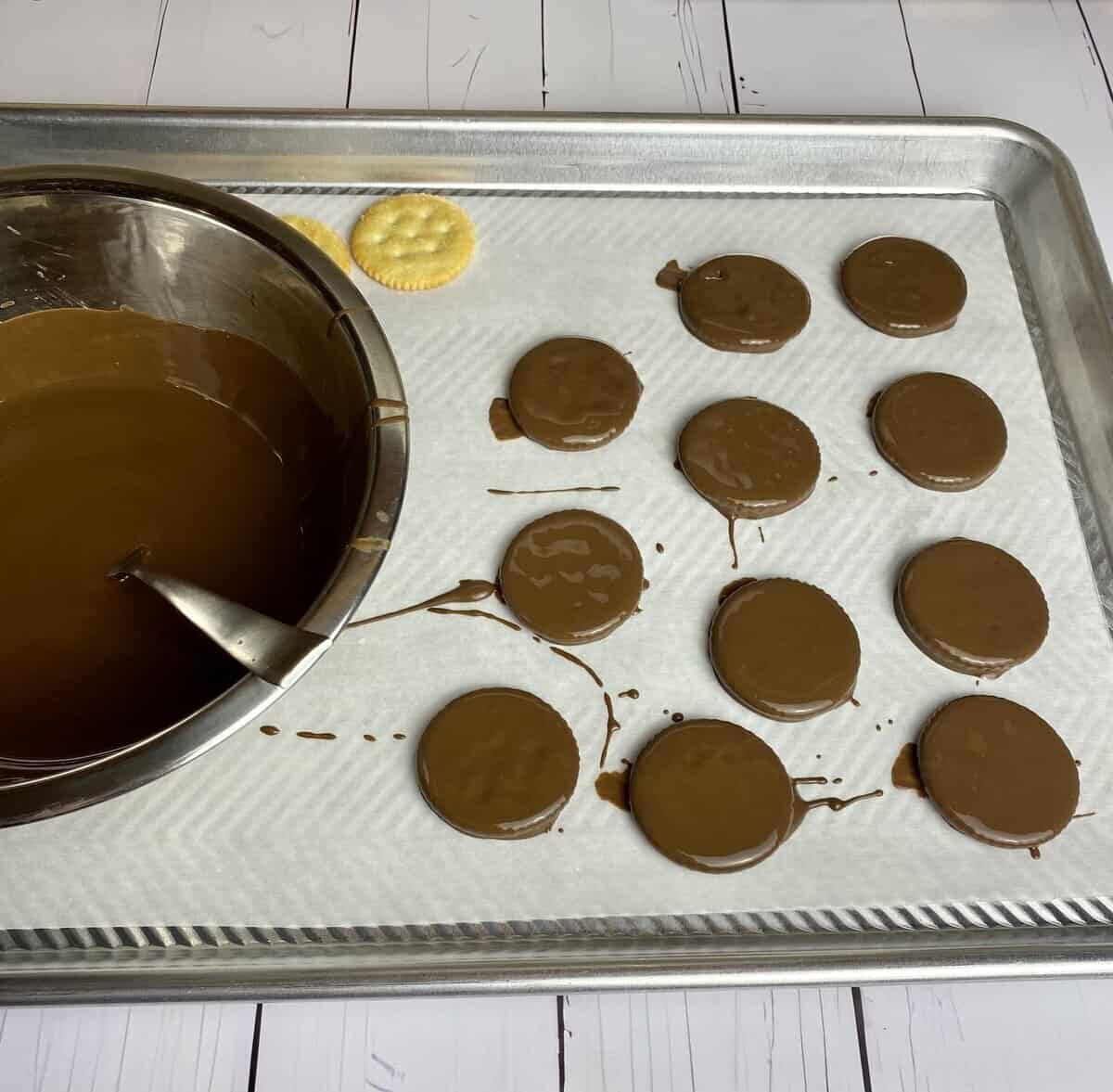 A bowl of melted andes chocolate on a sheet pan with parchment and chocolate dunked crackers cooling.