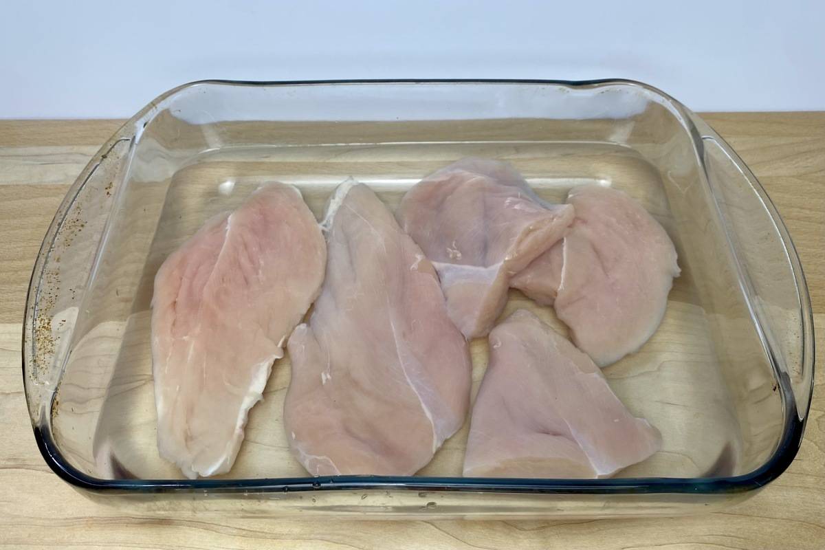 A glass casserole dish filled with saltwater brine and trimmed chicken breasts.