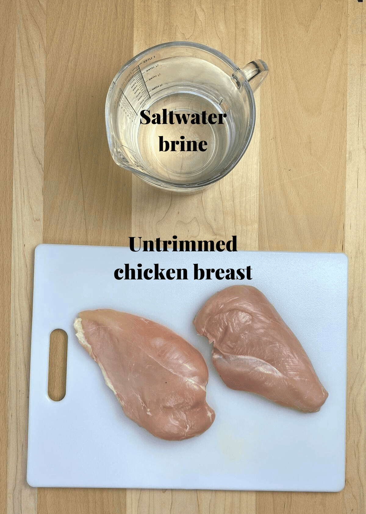 A cutting board with two chicken breasts next to a pitcher filled with saltwater brine.