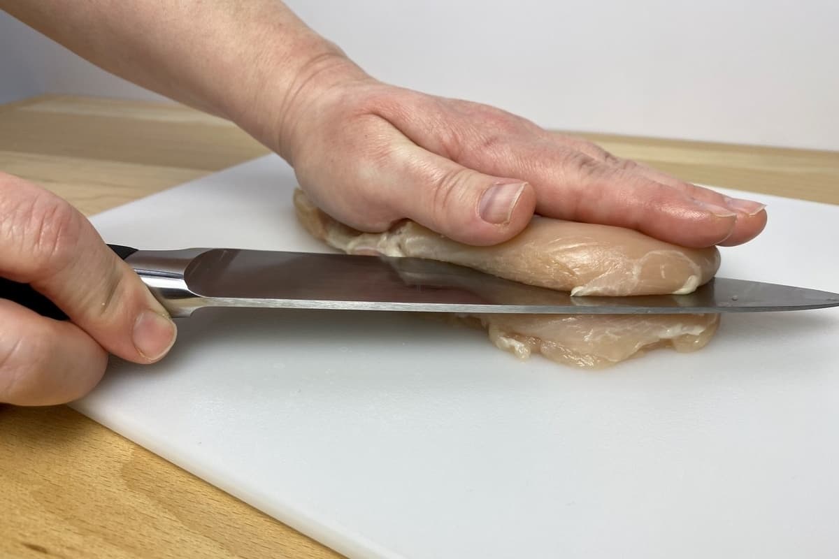 A hand holding a chefs knife slicing horizontally into a chicken breast.