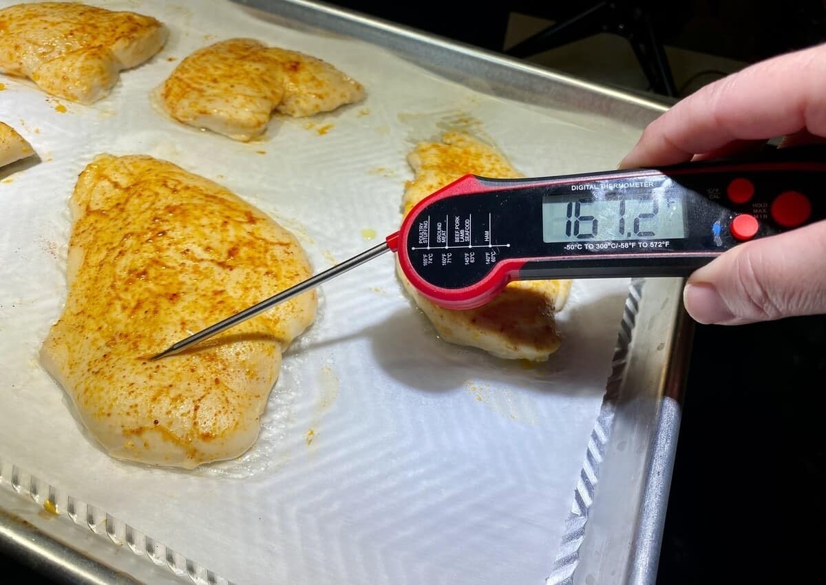 A thermometer checking the temperature of a piece of cooked chicken breast on a sheet pan.