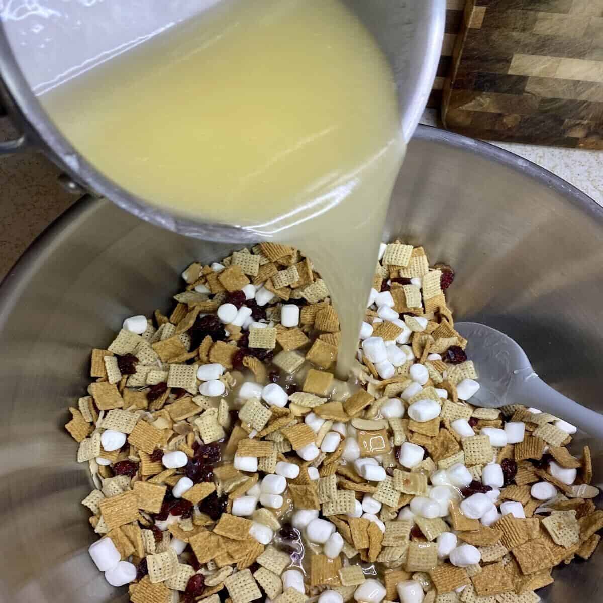 A small pot pouring the butter and sugar mix all over the cereal mixture in a large bowl.