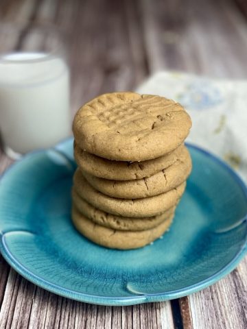 4 ingredient peanut butter cookies stacked up on a blue plate with a cup of milk alongside.