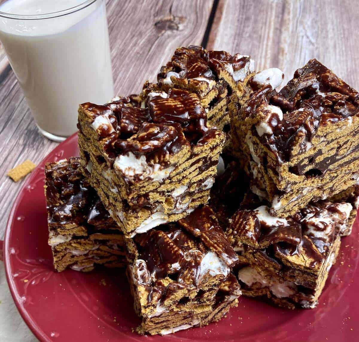 Red plate with smores bars cut into cubes and piled up next to a glass of milk.