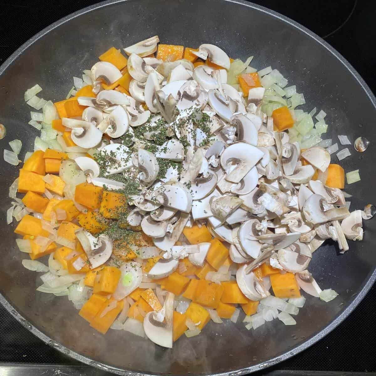 A saute pan cooking onions, yams, mushrooms, garlic, and rosemary.