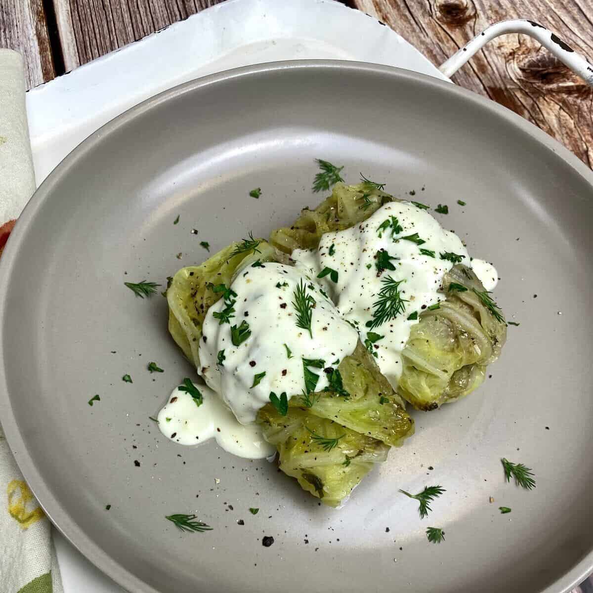 Two Greek cabbage rolls on a gray plate covered in lemon cream sauce and fresh herbs.