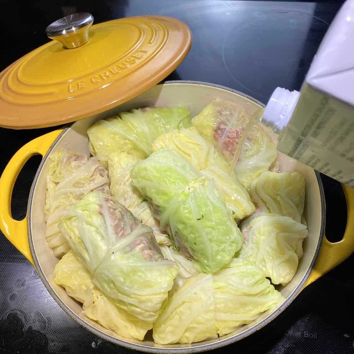 A pot filled with cabbage rolls and a box of chicken stock being poured in the pot.