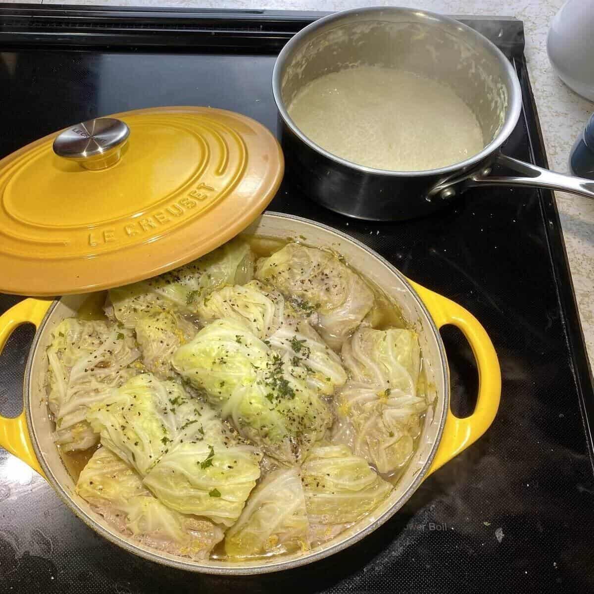 A pot filled with cooked cabbage rolls and a small pot reducing heaving whipping cream on the back burner.