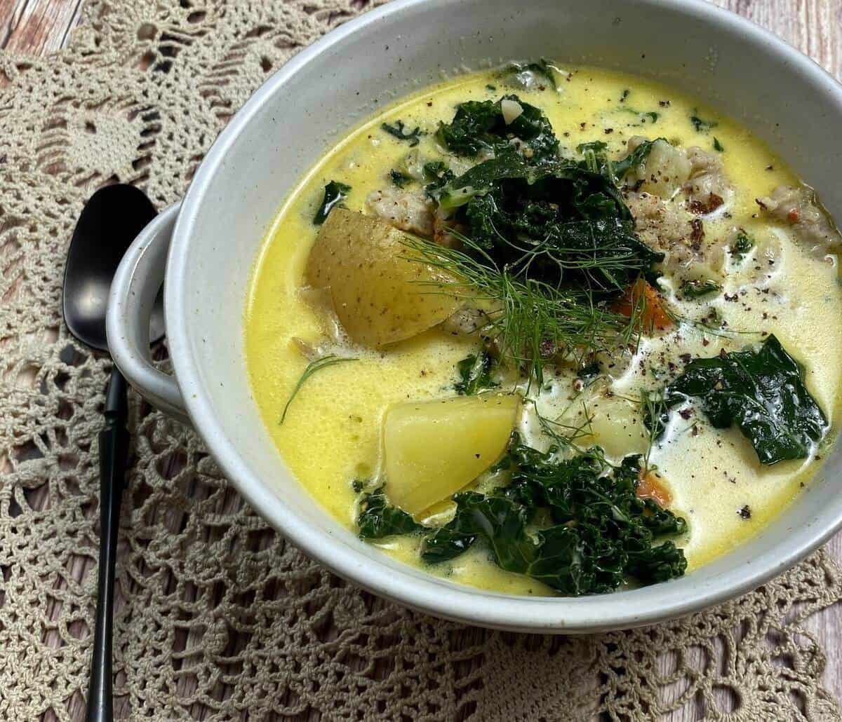 A closeup of a bowl of chicken kale and potato soup with a spoon and tablecloth alongside.