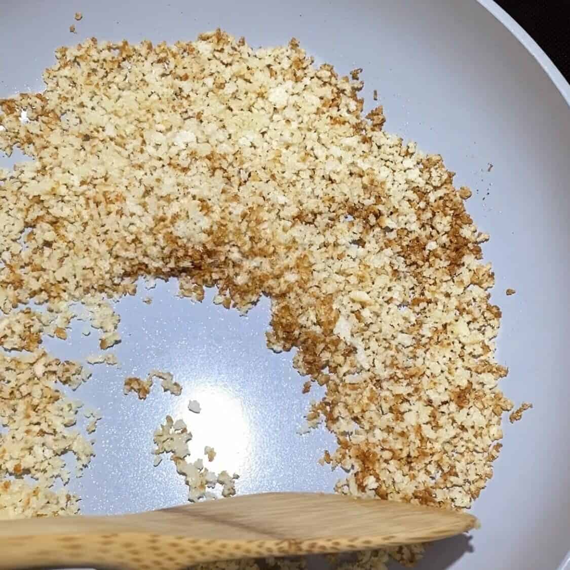 A skillet toasting panko breadcrumbs with a wooden spoon stirring.