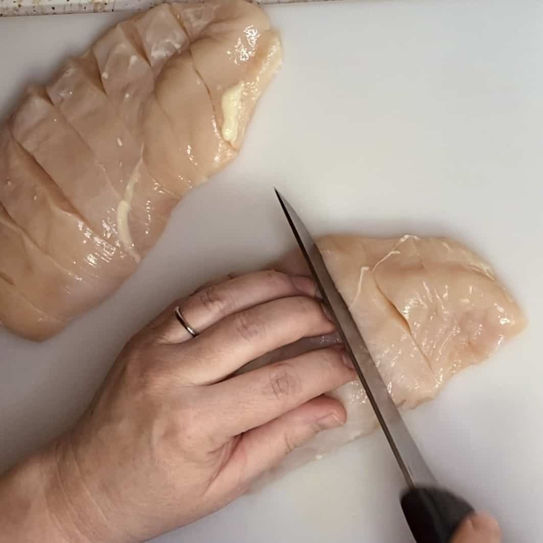 A hand on top of a raw chicken breast slicing with a knife.