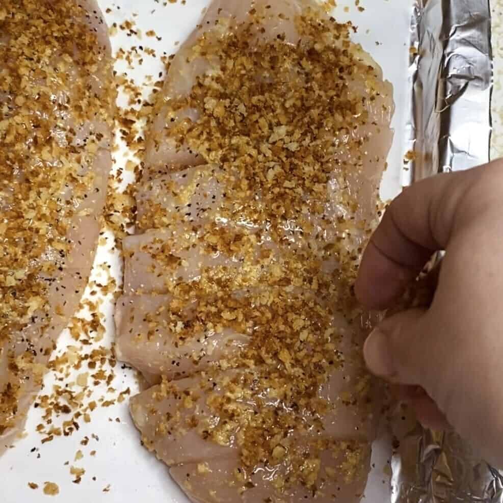 A hand sprinkling panko breadcrumbs on chicken breasts.