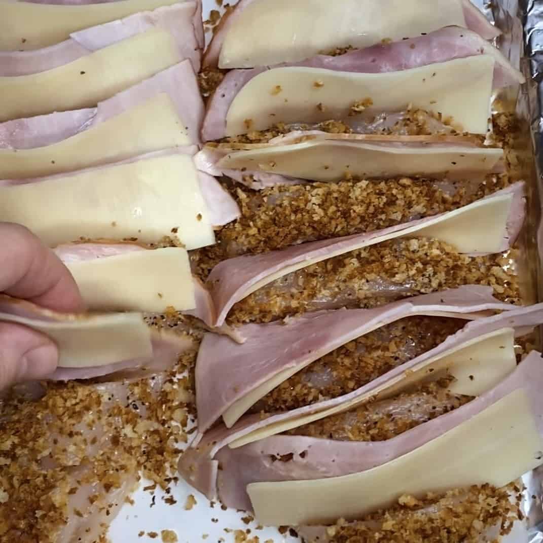 A hand stuffing ham and cheese into the slices of a hasselbacked chicken breast.