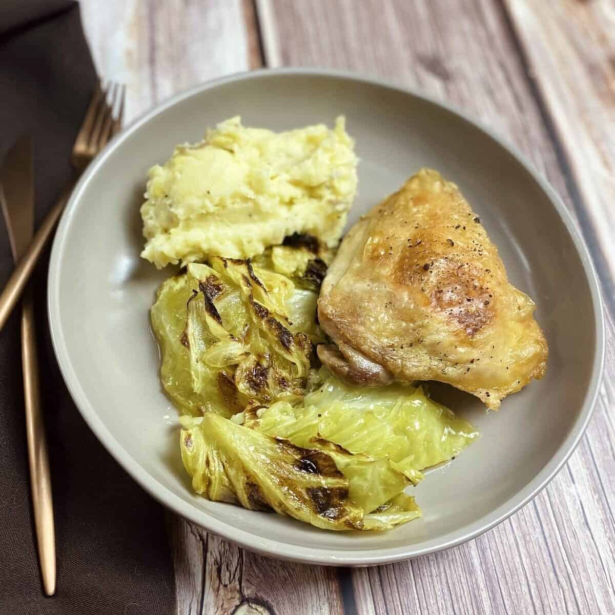  A plate with fried chicken thighs,  schmaltzy cabbage, and mashed potatoes alongside silverware.