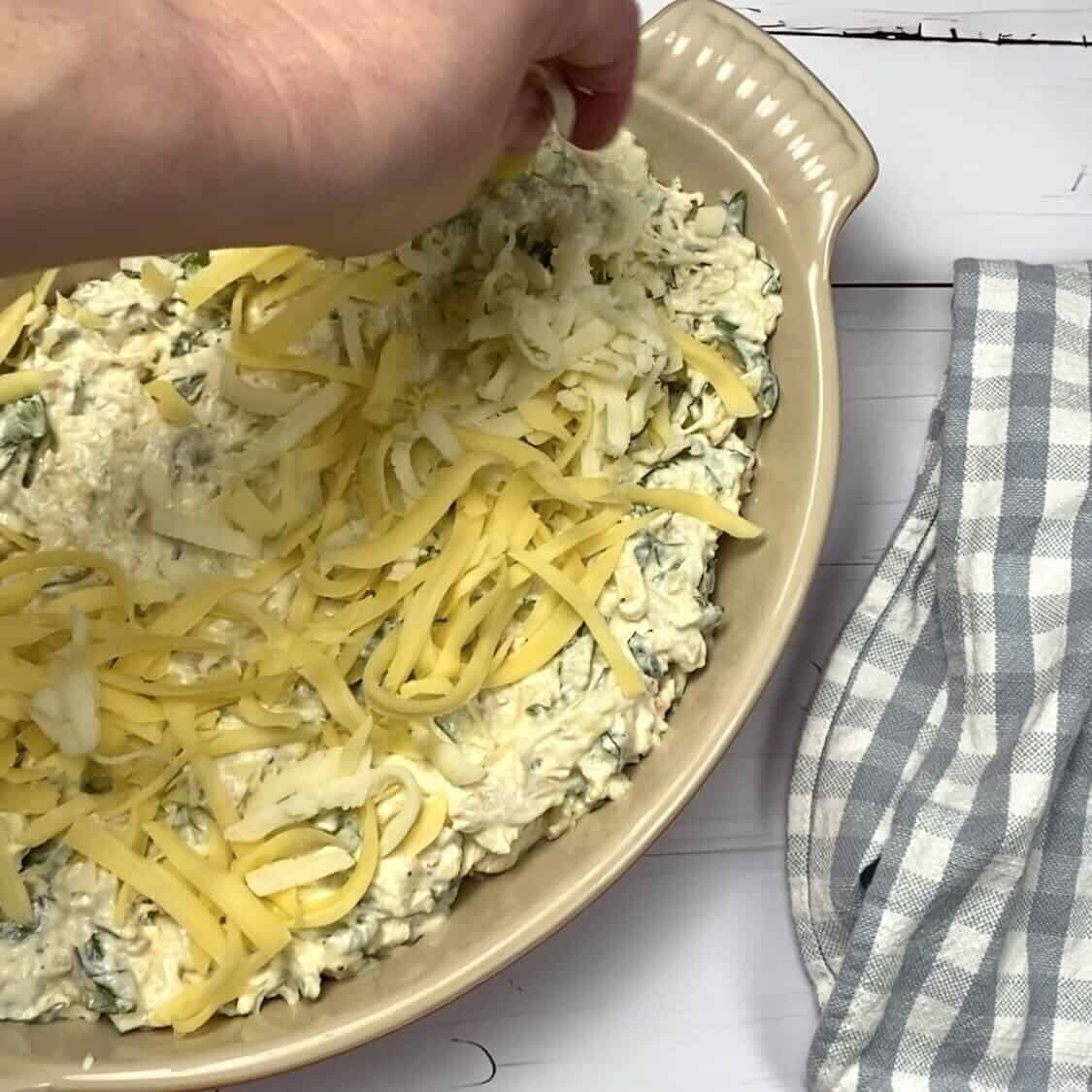 A hand adding shredded cheese to the top of a dish of crab and spinach dip.