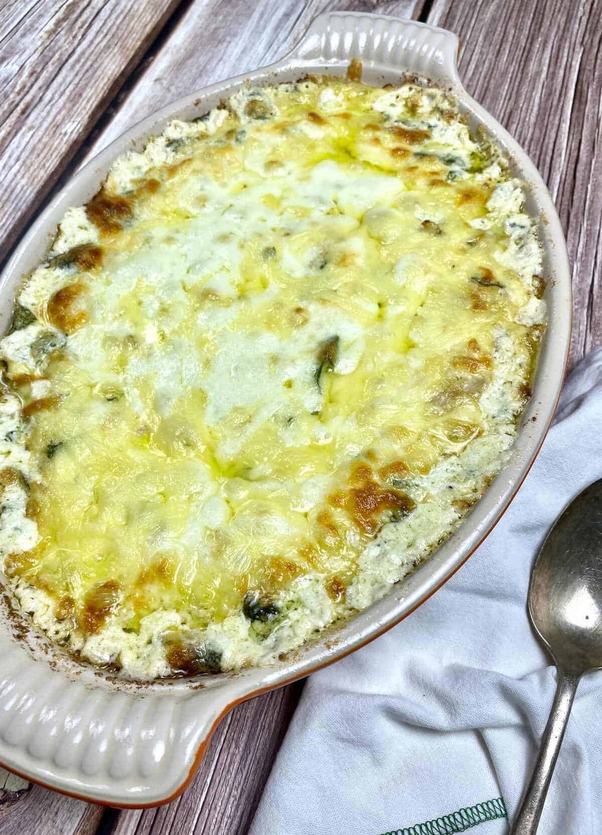 A casserole dish with baked crab and spinach dip alongside a serving spoon.