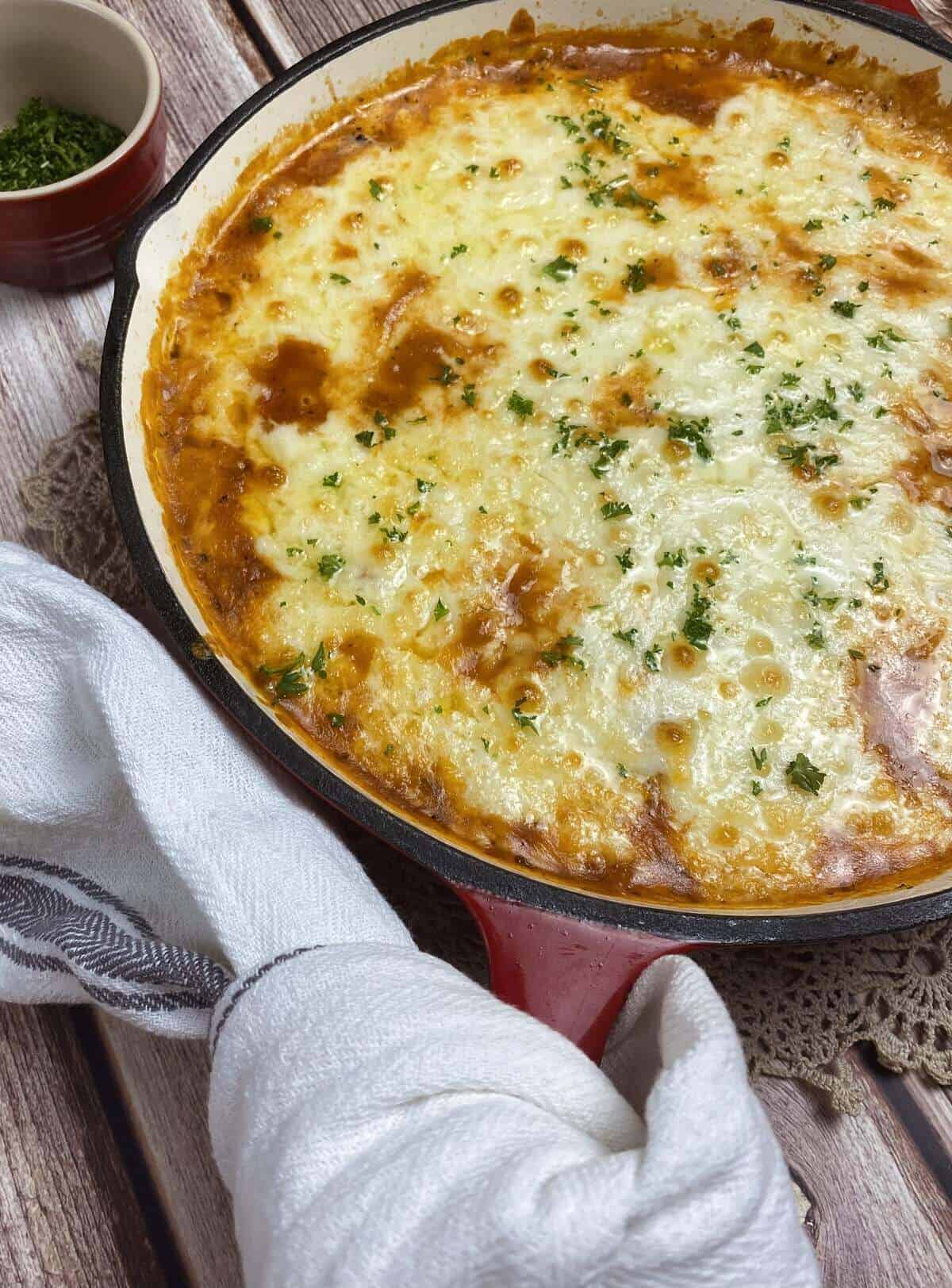 A close up of the skillet with baked cheesy white bean bake with a towel and parsley.