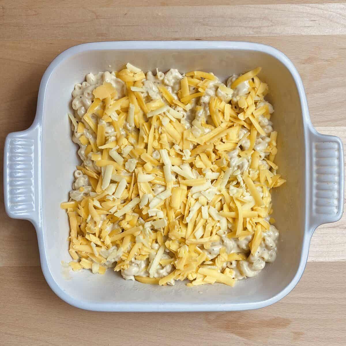 A casserole dish with half the cheesy pasta and more shredded cheese on top.