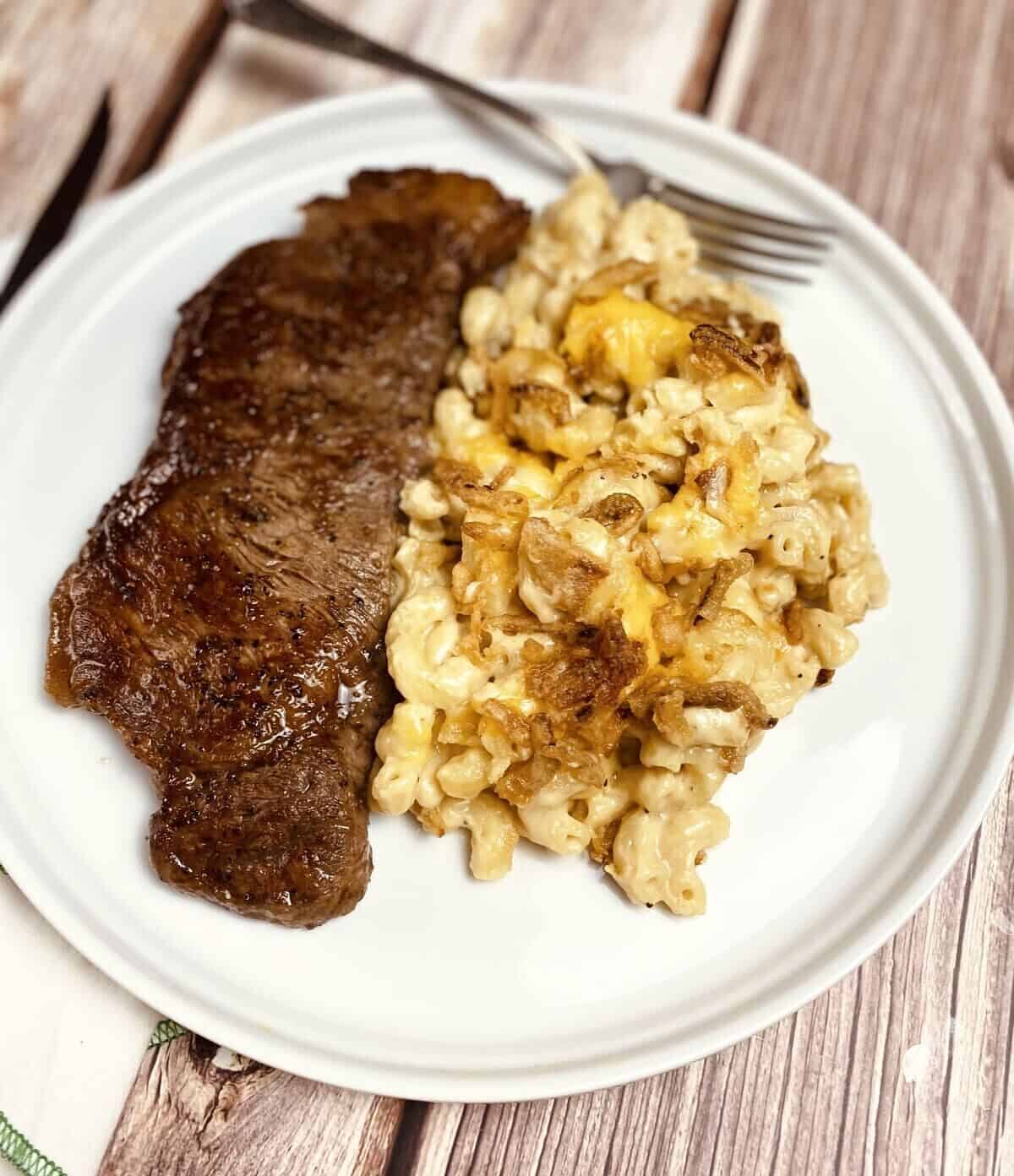 A white plate with a pile of cheesy mac, a steak, and a fork.