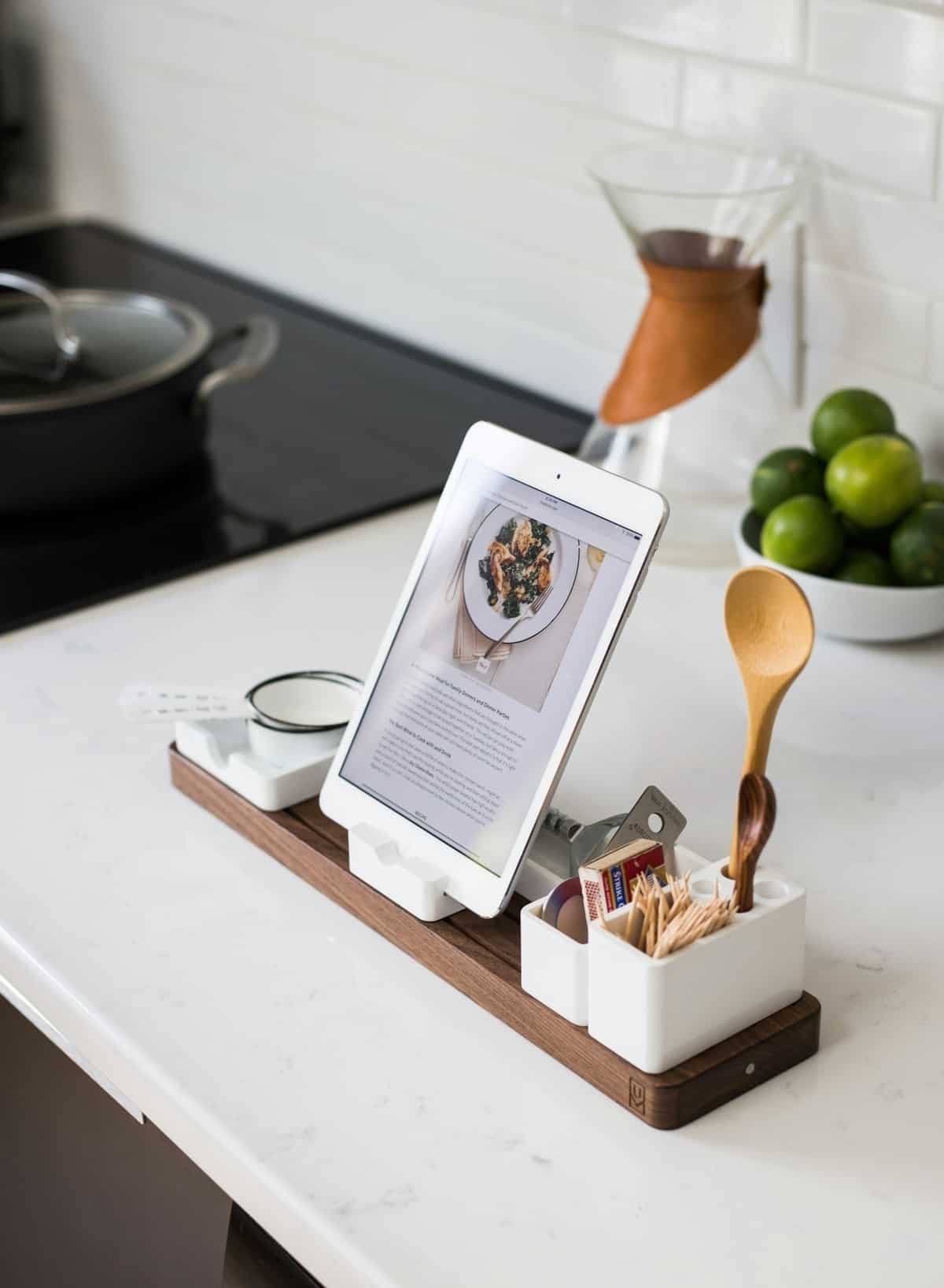 A kitchen counter with a tray holding an ipad with a recipe along with measuring cups, a wooden spoon and other tools.