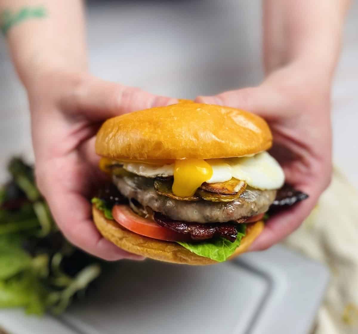 Two hands squishing a burger together with runny egg yolk pouring out.