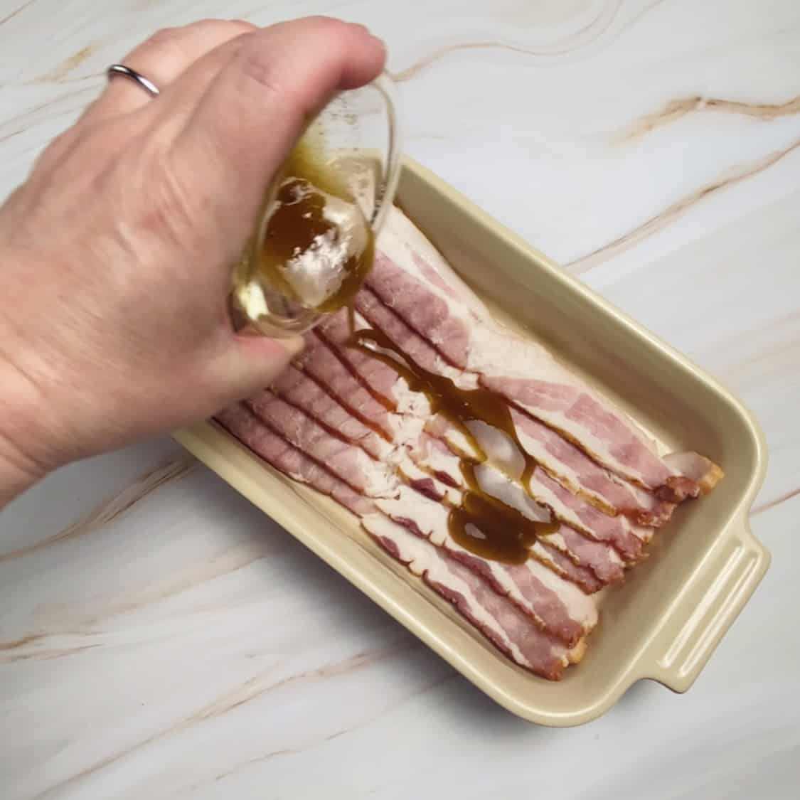 A hand pouring the maple and brown sugar mix onto the tray of bacon.