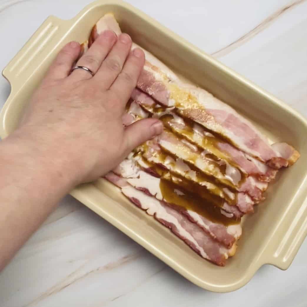 A hand rubbing maple brown sugar mix all over a tray of bacon.