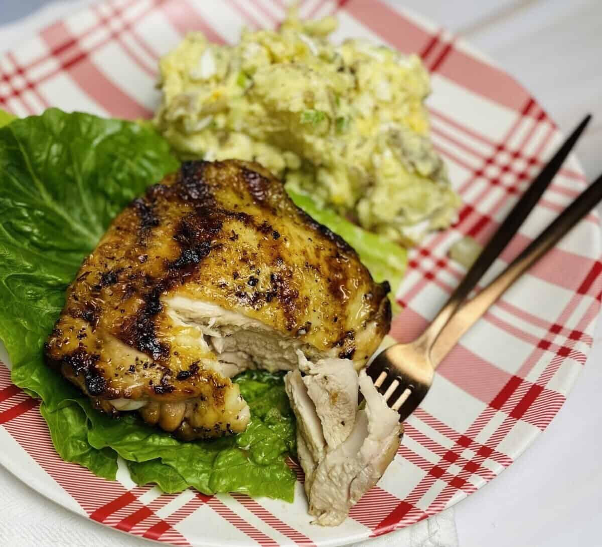 A piece of grilled chicken on a plate with lettuce, potato salad, and silverware.