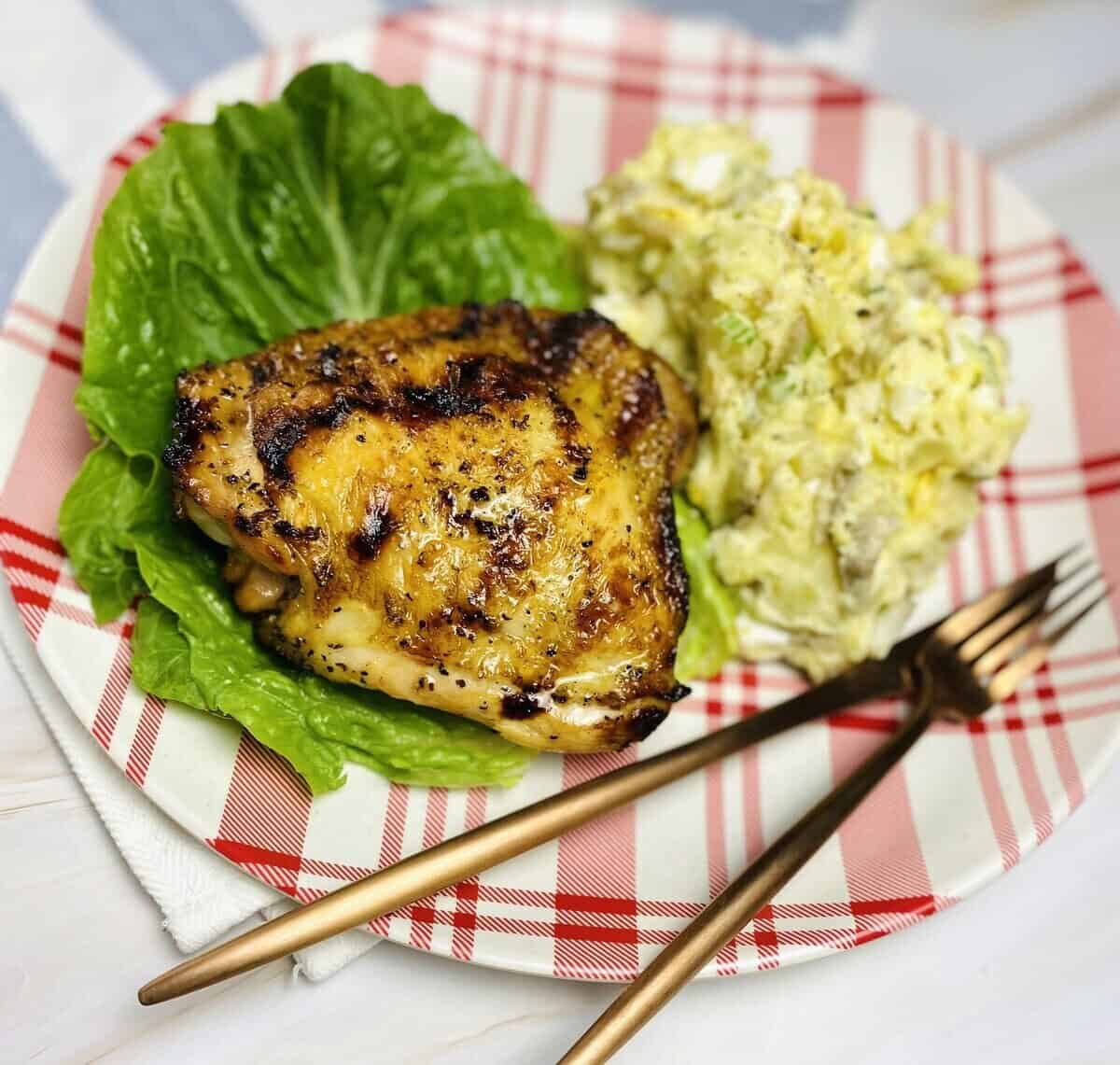 Grilled chicken on a plate with potato salad, lettuce, and silverware.