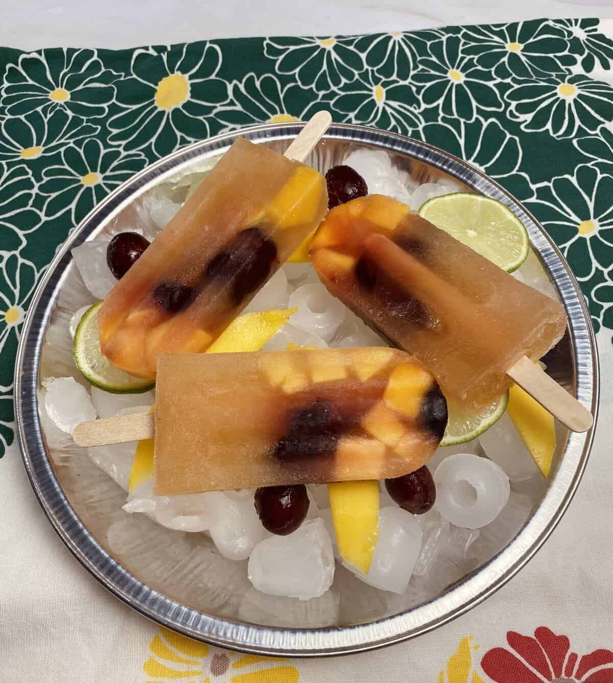 Three popsicles on a round tray of ice with fresh fruit.