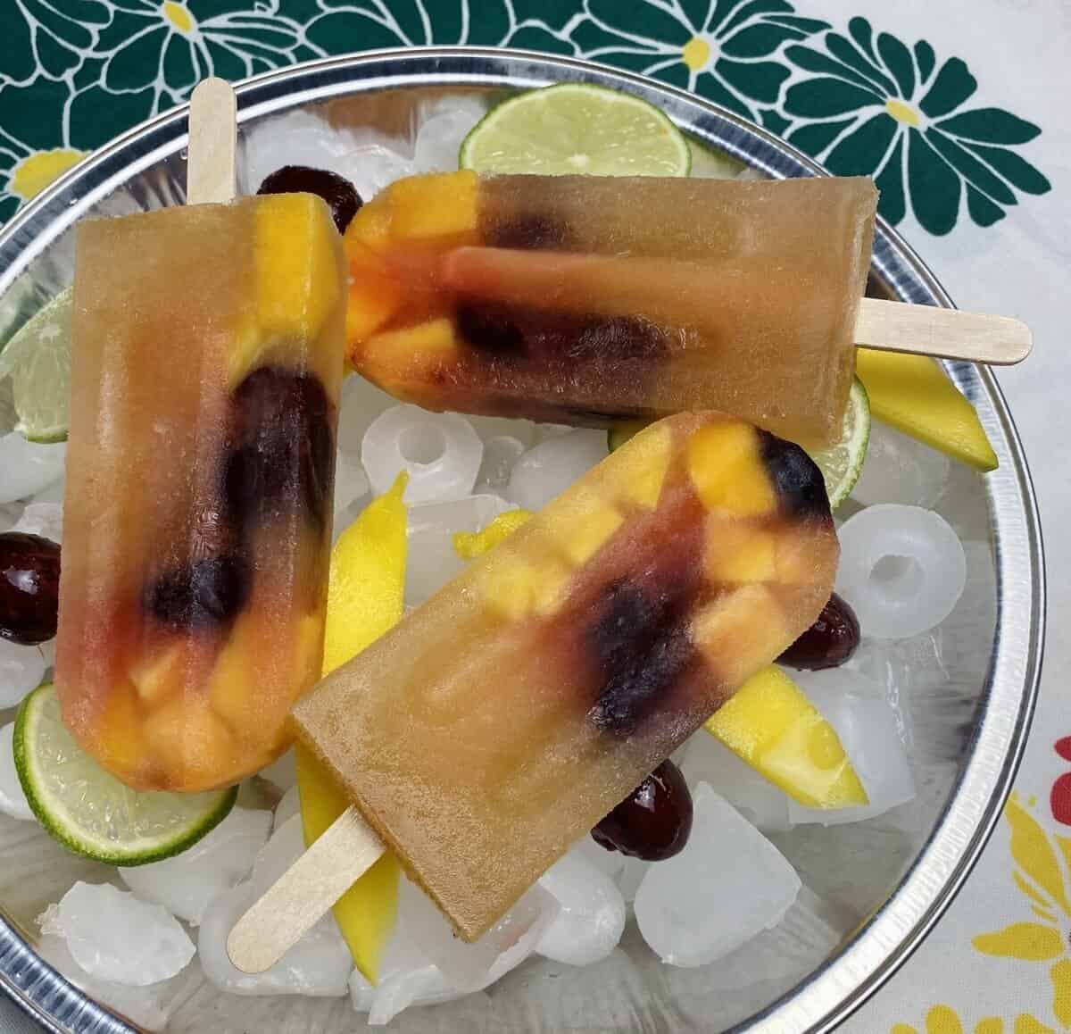 Three white sangria popsicles on a tray of ice garnished with fresh fruit.