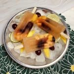 Three white sangria popsicles sitting on a tray of ice garnished with fresh fruit.