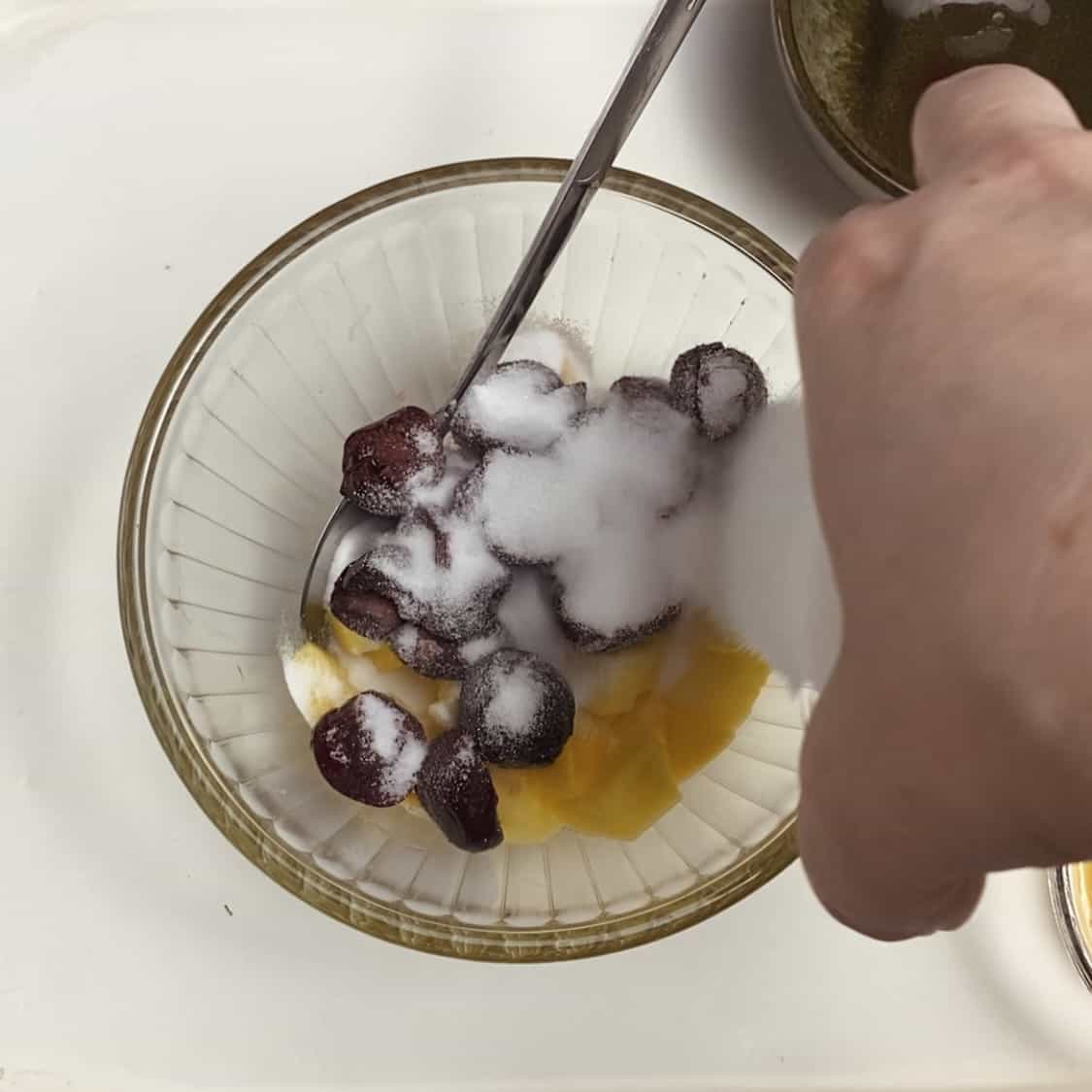 A hand pouring sugar into a bowl of fruit.