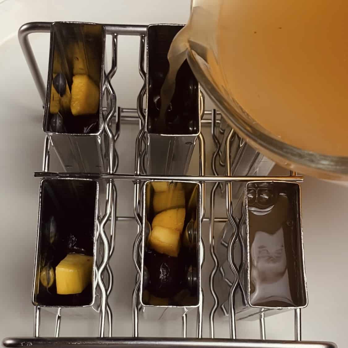 Punch being poured into metal popsicle molds that have chopped fruit in them.