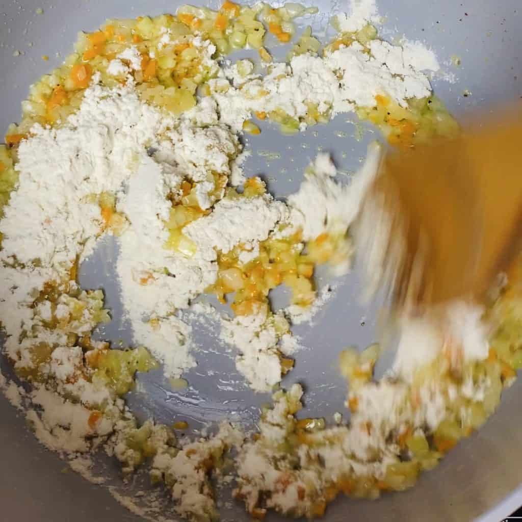 Flour being stirred into sauted mire poix in a pot.