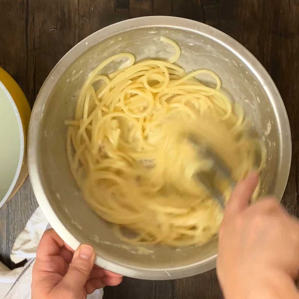 A hand with tongs stirring pasta and cheese together.