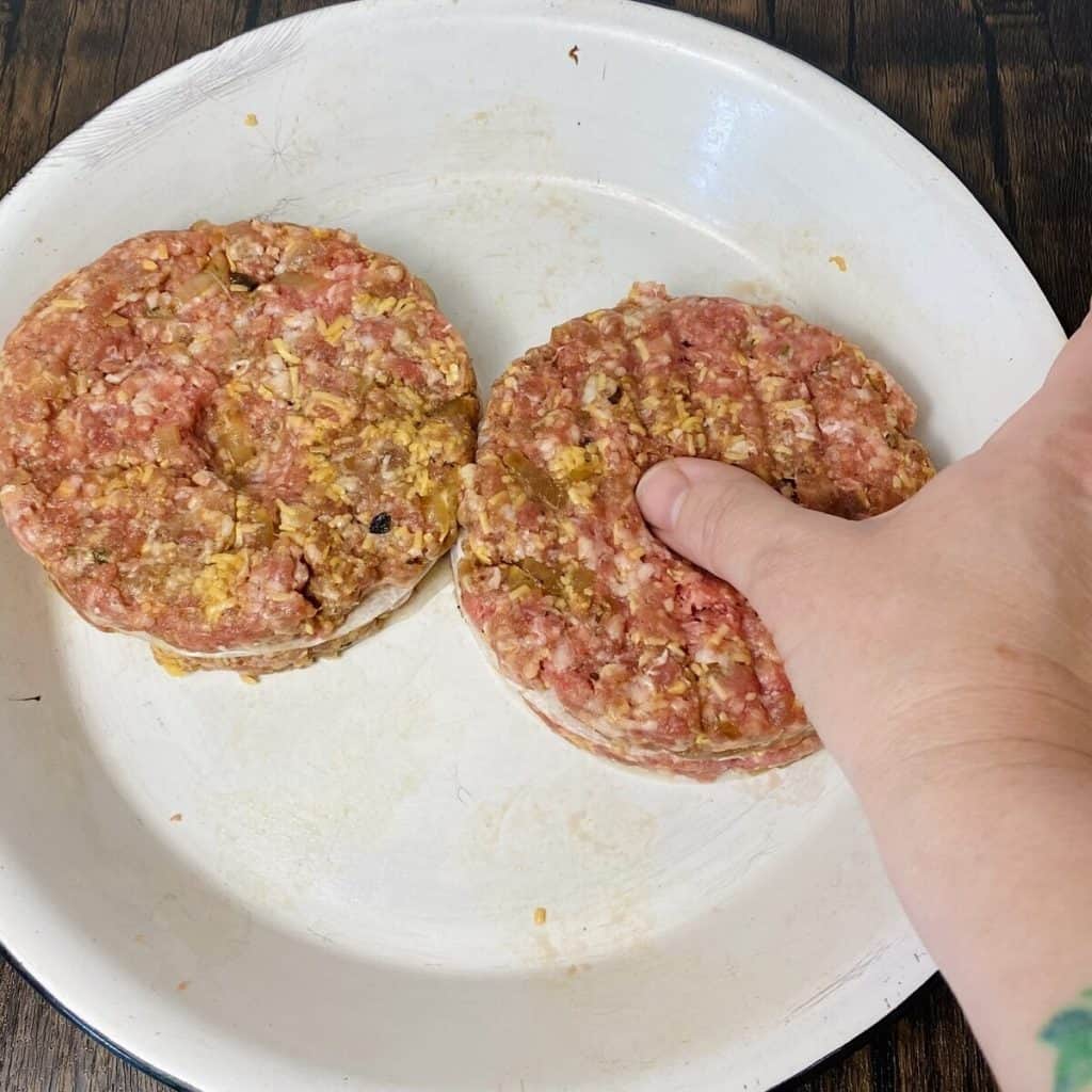 A thumb pressing a dent into the top of a raw burger patty.