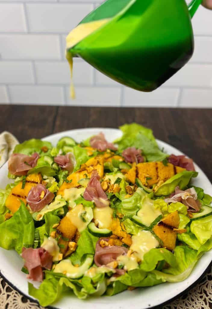 A green pitcher pouring dressing onto the grilled melon and prosciutto salad.